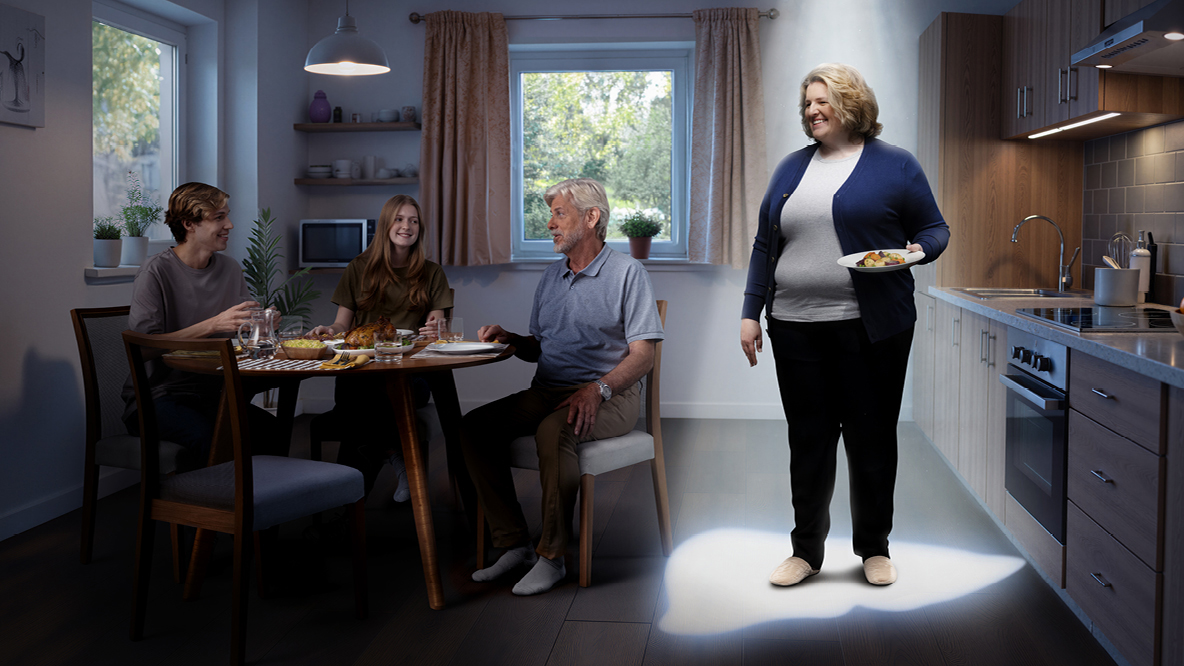 Adipöser Mensch steht in schattiger Küche, beleuchtet mit einem Lichtkegel in Leberform.